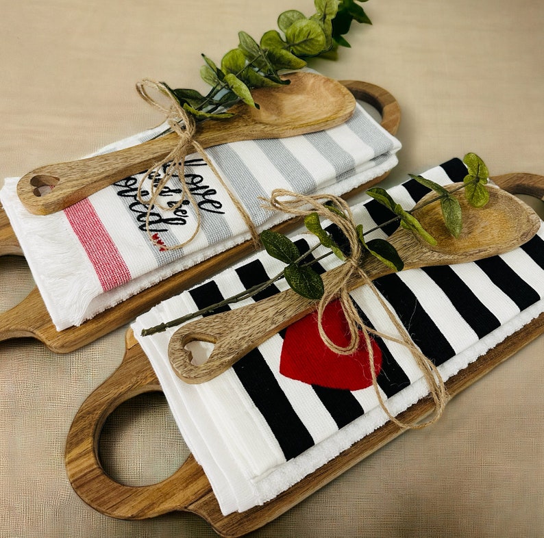 May include: Two wooden cutting boards with handles, each with a white kitchen towel, a wooden spoon, and twine. One towel has red and white stripes and says "I love you more than bread". The other towel has black and white stripes and a red heart.