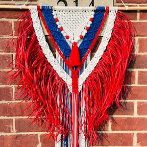 May include: A red, white, and blue macrame wall hanging with fringe. The macrame is in the shape of a triangle with a beaded string hanging from the top.