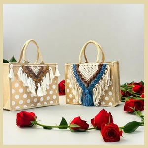 May include: Two beige jute tote bags with white polka dots and decorative macrame trim. The bags have white and brown tassels and a blue tassel. The bags are sitting on a table with red roses.