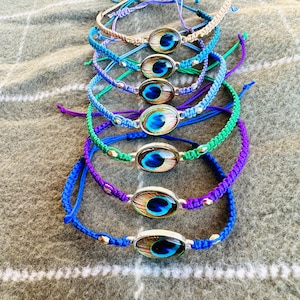 May include: A collection of handmade bracelets with a peacock feather design. Each bracelet features a woven cord in various colors, including blue, green, and purple, and a silver-toned oval centerpiece. The bracelets are displayed on a gray surface.