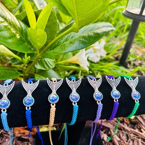 May include: A set of eight mermaid tail charm bracelets with iridescent blue and green stones. The bracelets are made with colorful braided cords and feature silver mermaid tail charms.