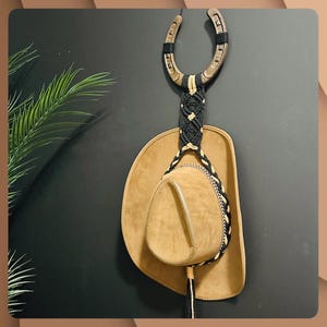 May include: A brown cowboy hat hangs from a horseshoe-shaped wall mount. The horseshoe is made of wood and black accents. A black and tan macrame cord connects the horseshoe to the hat. The hat is tan with a decorative band.