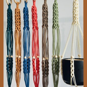 May include: A collection of macrame plant hangers in various colors, including teal, orange, blue, red, brown, and green. Each hanger features a wooden ring at the top and wooden beads. One hanger holds a black ceramic pot.