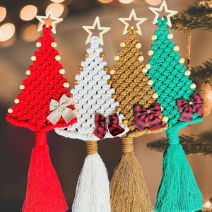 May include: Four macrame Christmas tree ornaments in red, white, gold, and teal. Each tree has a wooden star topper, wooden bead accents, and a decorative bow. The trees have tassels at the bottom, perfect for holiday decor.