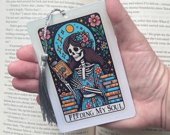 May include: A white bookmark featuring a colourful illustration of a skeleton holding a book. The skeleton is wearing a blue dress with floral patterns. The bookmark has a silver tassel and the text "Feeding My Soul".