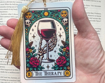 May include: A tarot card bookmark with a white background and a colourful illustration of a skeleton hand holding a glass of red wine. The card is labelled "The Therapy" and has the number "III" and a circle with a symbol inside. The bookmark has a gold tassel.
