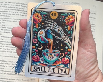 May include: A silver bookmark with a colorful illustration of a skeleton pouring tea from a cup. The bookmark has the text "Spill the Tea" and the number "411" in a circle. The bookmark has a blue tassel.