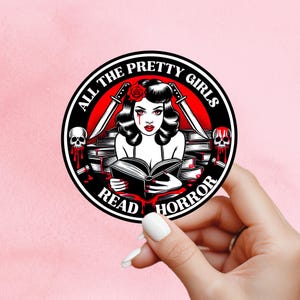 May include: A circular sticker featuring a vintage-style illustration of a woman reading a book, with knives and skulls. The text reads "ALL THE PRETTY GIRLS READ HORROR". The design uses red, black, and white colours.