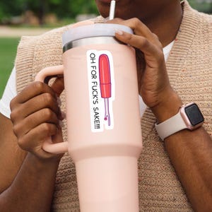 May include: A pink insulated tumbler with a white lid and handle. A sticker on the tumbler features a red and pink illustration of a tool with the text "OH FOR FUCK'S SAKE!!!". The person is wearing a beige sweater and a rose gold watch.