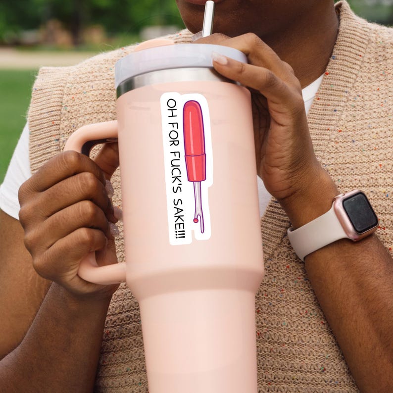 May include: A pink insulated tumbler with a white lid and handle. A sticker on the tumbler features a red and pink illustration of a tool with the text "OH FOR FUCK'S SAKE!!!". The person is wearing a beige sweater and a rose gold watch.