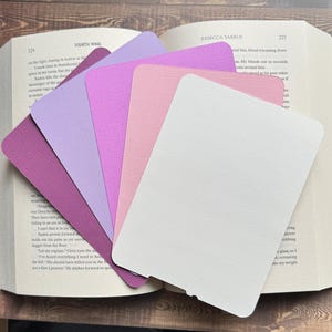 May include: An open book with pages marked by a fan of colorful, textured paper bookmarks. The bookmarks are in shades of purple, pink, and white. The book is titled "Fourth Wing" and "Rebecca Yarros".