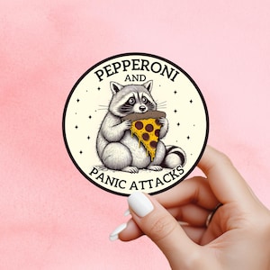 May include: A circular sticker with a cartoon raccoon eating pizza. The text "PEPPERONI AND PANIC ATTACKS" is printed around the raccoon. The raccoon is grey and white, and the pizza slice has yellow cheese and red pepperoni. The sticker is held by a hand.