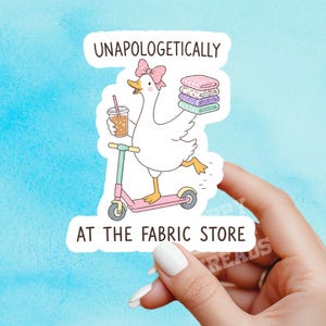 May include: A die-cut sticker featuring a white cartoon duck wearing a pink polka-dot bow, riding a pink and teal scooter while holding an iced coffee and a stack of folded fabric. The text reads UNAPOLOGETICALLY AT THE FABRIC STORE.