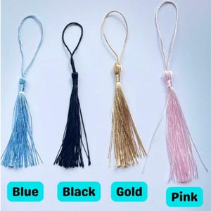 May include: Four tassels in blue, black, gold, and pink, each with a looped cord for hanging. The colors are labeled below each tassel. The tassels are made of a silky material.