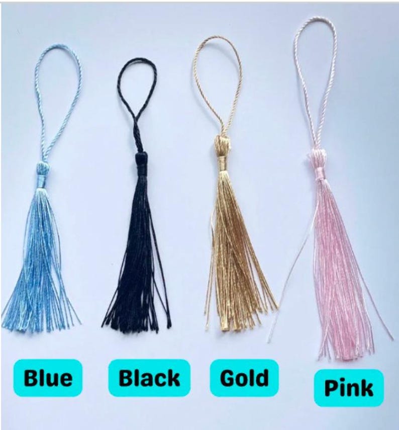 May include: Four tassels in blue, black, gold, and pink, each with a looped cord for hanging. The colors are labeled below each tassel. The tassels are made of a silky material.
