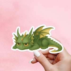 May include: A green dragon sticker with yellow detailing, showing a grumpy expression. The sticker has a white border and is held by a hand against a pink background. The dragon has yellow eyes and wings.