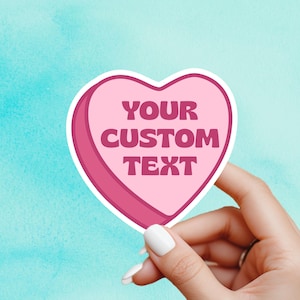 May include: A pink heart-shaped sticker with the text "YOUR CUSTOM TEXT" in dark pink letters. The sticker is held by a hand with white nail polish against a teal background.