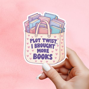 May include: A sticker featuring a tote bag filled with books in various colors. The bag has the text "PLOT TWIST... I BROUGHT MORE BOOKS" and is decorated with pink hearts. The sticker is held by a hand with white nail polish.
