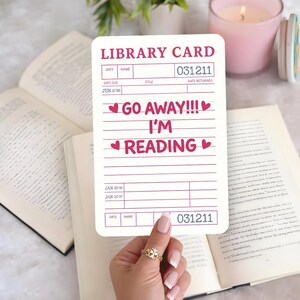 May include: A white library card with pink text saying "GO AWAY!!! I'M READING" and heart symbols. The card has spaces for details and a library number. The card is held over an open book.