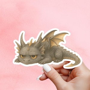 May include: A sticker of a grumpy cartoon dragon in grey, brown, and gold tones. The dragon has horns, wings, and a long tail, and is lying down. The sticker is held by a hand against a pink background.