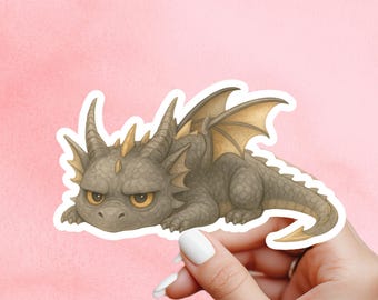 May include: A sticker of a grumpy cartoon dragon in grey, brown, and gold tones. The dragon has horns, wings, and a long tail, and is lying down. The sticker is held by a hand against a pink background.