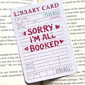 Può includere: Un adesivo per la tessera della biblioteca con la scritta "SORRY I'M ALL BOOKED" in rosso, con accenti a cuore. La tessera ha un design vintage con linee per data, nome e titolo, e il numero 031211. L'adesivo è su un libro aperto.