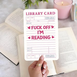 May include: A white library card with pink text that reads "FUCK OFF I'M READING" with heart symbols. The card also has the words "LIBRARY CARD" and other fields. The card is held over an open book.