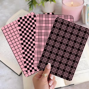 May include: Four bookmarks with pink and black patterns are displayed over an open book. The designs include a grid, a checkerboard, and a plaid. A hand with a gold ring holds the bookmarks. A lit pink candle and a small plant are in the background.