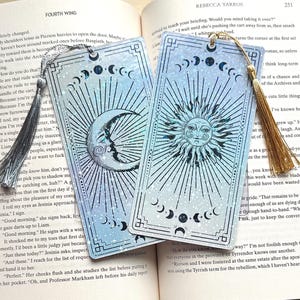 May include: Two celestial-themed bookmarks with silver tassels. One features a crescent moon with a face, the other a sun with a face. Both have a blue and silver glitter effect and a border.