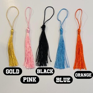 May include: Five colorful tassels in gold, pink, black, blue, and orange. Each tassel has a loop at the top and a bundle of threads at the bottom. The colors are labeled with text: GOLD, PINK, BLACK, BLUE, and ORANGE.