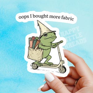 May include: A sticker depicting a green frog on a scooter, wearing a hat, and carrying a bag filled with fabric rolls. The text "oops I bought more fabric" is above the frog. The background is a light blue watercolour wash.