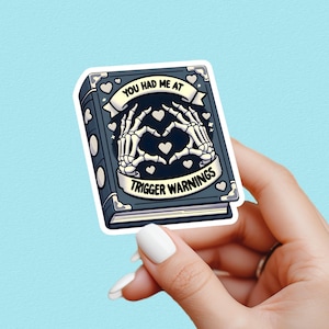 May include: A sticker with a dark blue book design and the text "YOU HAD ME AT TRIGGER WARNINGS." The cover shows skeleton hands forming a heart shape. White hearts and stars decorate the design. The sticker has a white border.