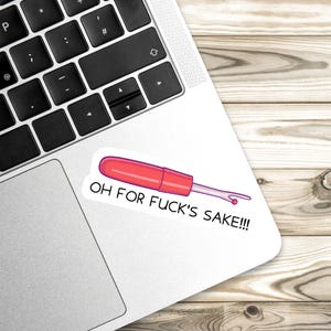 May include: A sticker featuring a red seam ripper with the text "OH FOR FUCK'S SAKE!!!" in black. The sticker is on a white surface, next to a laptop keyboard, with a wooden background.