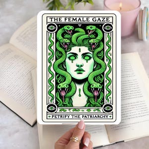 May include: A white sticker with a Medusa illustration. The image shows a woman with green skin and snakes for hair. The text "THE FEMALE GAZE" is at the top and "PETRIFY THE PATRIARCHY" at the bottom.