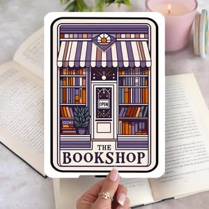 May include: A sticker depicting a bookstore with a striped awning, open door, and shelves of books. The text "THE BOOKSHOP" is at the bottom. The sticker is held over an open book.