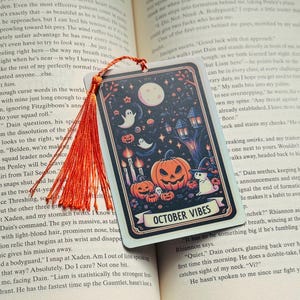 May include: A rectangular Halloween-themed bookmark with a vibrant illustration. The design features a full moon, pumpkins, ghosts, and the text "OCTOBER VIBES". An orange tassel hangs from the top.