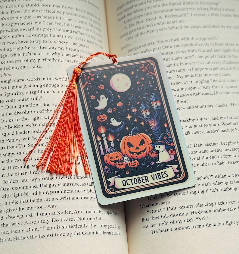 May include: A rectangular Halloween-themed bookmark with a vibrant illustration. The design features a full moon, pumpkins, ghosts, and the text "OCTOBER VIBES". An orange tassel hangs from the top.