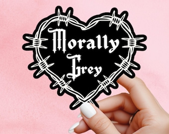 May include: A black heart-shaped sticker with the words "Morally Grey" in white gothic font, encircled by a white barbed wire design. The sticker is held against a pink background.