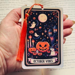 May include: A Halloween-themed bookmark featuring a vibrant illustration of pumpkins, ghosts, and a full moon against a dark night sky. The bookmark has an orange tassel and the words "OCTOBER VIBES" on a banner.