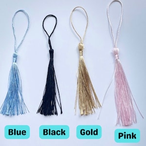 Op de afbeelding: Vier kwast sleutelhangers in verschillende kleuren: blauw, zwart, goud en roze. De kwasten zijn gemaakt van dunne, zachte draden.