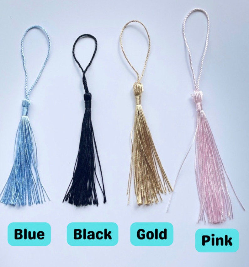 Op de afbeelding: Vier kwast sleutelhangers in verschillende kleuren: blauw, zwart, goud en roze. De kwasten zijn gemaakt van dunne, zachte draden.