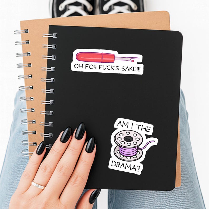 May include: A black notebook with two stickers: one with a red and white sewing tool and the text "OH FOR FUCK'S SAKE!!!", and another with a pink spool of thread and the text "AM I THE DRAMA?". The notebook is on top of a brown notebook.