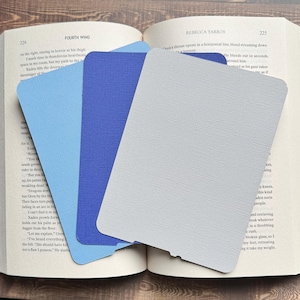 May include: Three textured paper bookmarks in shades of blue and grey rest on an open book. The bookmarks are rectangular with rounded corners, and one has a notched edge. The book pages display text from a novel.