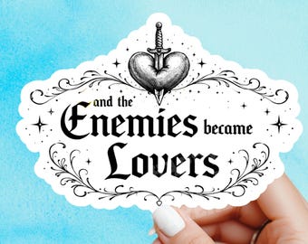 May include: A white sticker with the text "and the Enemies became Lovers" in a decorative font. The sticker features a heart pierced by a sword, surrounded by stars and flourishes, held by a hand against a blue background.