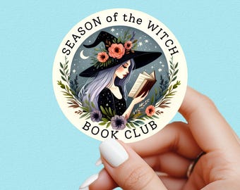 May include: A round sticker with a witch reading a book, surrounded by floral and celestial elements. The sticker reads "SEASON of the WITCH BOOK CLUB" in a circular design. The witch wears a black hat with flowers.