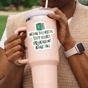 Pu&ograve; includere: Un grande bicchiere termico rosa chiaro con coperchio e manico bianchi. Un adesivo sul bicchiere presenta il logo di Microsoft Excel e il testo "When You Excel They Always Spreadsheet About You" in verde, viola e bianco.