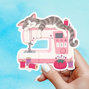 May include: A die-cut sticker featuring a grey tabby cat sleeping on top of a light pink sewing machine. The illustration includes several spools of pink and purple thread and a round red pincushion with pins at the base of the machine.