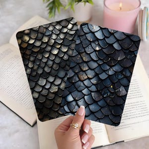 May include: Two square coasters with a dark, scale-like pattern, held over an open book. The coasters have a metallic sheen with hints of gold and blue. The background includes a lit candle and a small plant.
