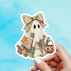 May include: A die-cut sticker featuring a ghost draped in a patchwork quilt with various floral and plaid patterns in muted earth tones. The ghost wears a large matching patchwork bow on its head and carries a small orange jack-o'-lantern bucket.