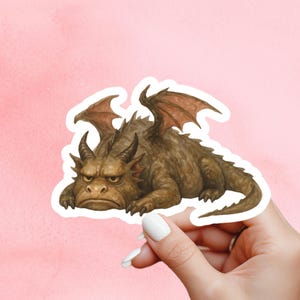 May include: A brown dragon sticker with a grumpy expression, horns, and wings. The sticker is on a white background, held by a hand with white nail polish, against a pink background. The dragon is lying down.